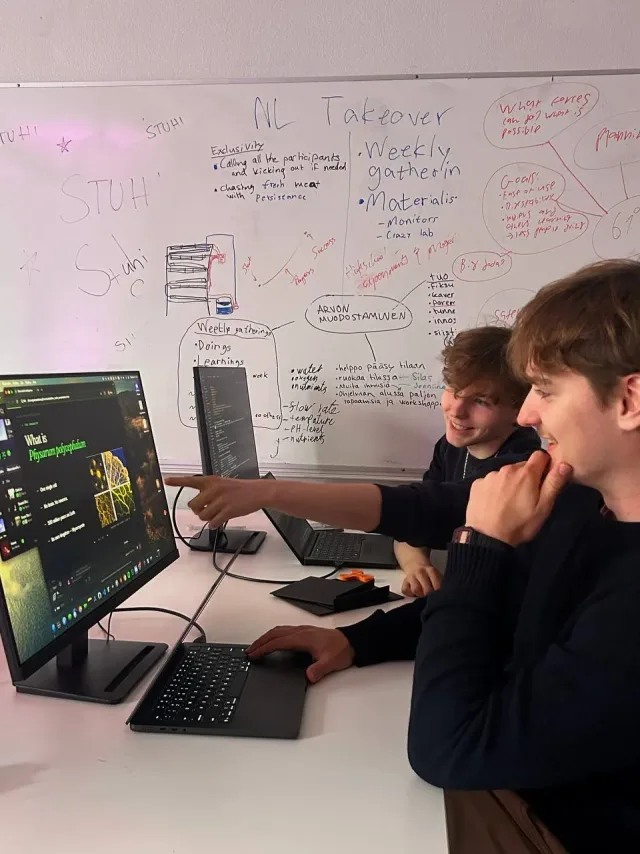Two students at a bank of screens in a STUHI workspace, a whiteboard full of notes behind them