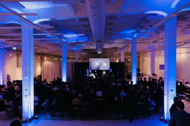 A packed hall lit blue at STUHI's Slush side event, a speaker on the screen at the far end